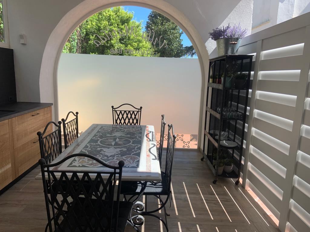 A covered dining area with wrought iron furniture opens onto a terrace with views of lush green trees and blue sky.