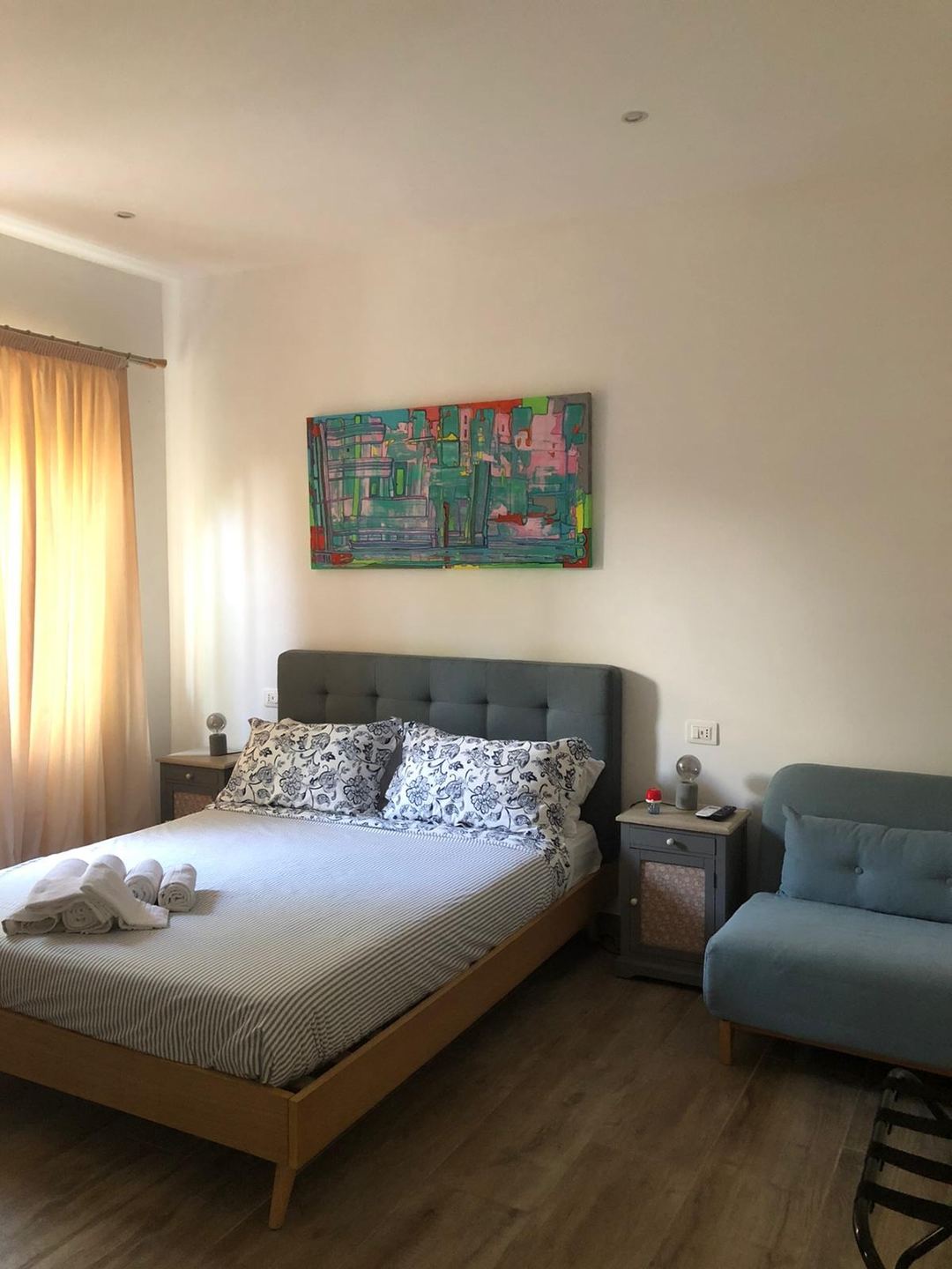 A bright bedroom at Villa Matilde B&B featuring a wooden bed with gray upholstered headboard, patterned pillows, white rolled towels, a colorful abstract painting, and natural light from yellow curtains.