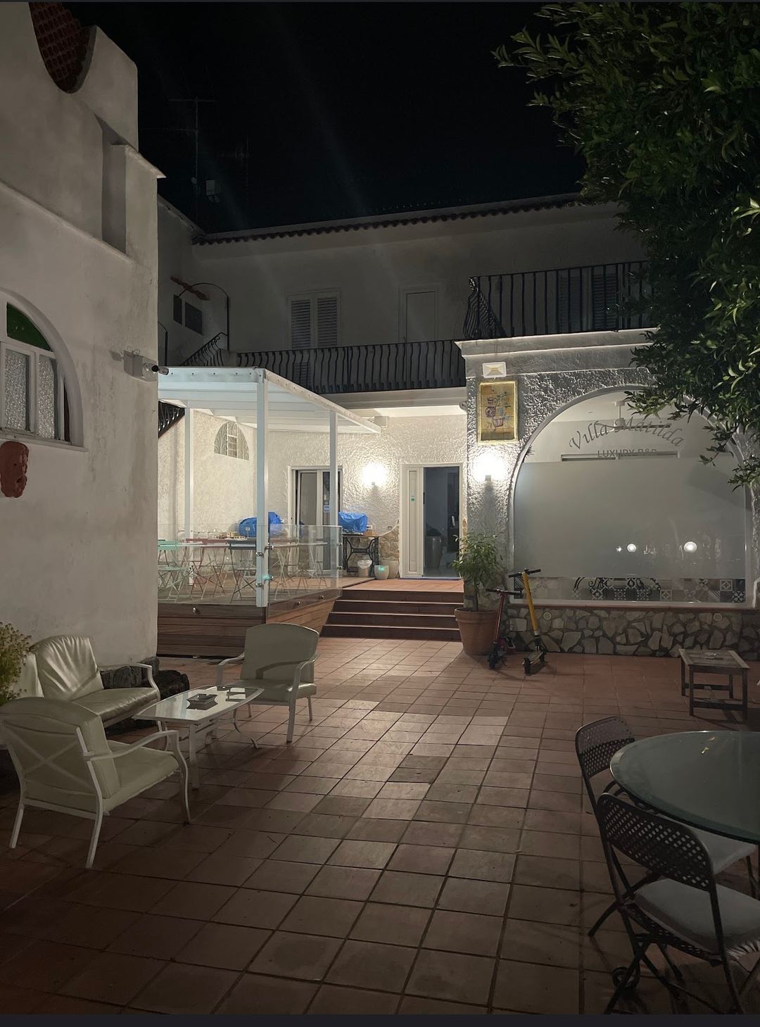 Illuminated nighttime courtyard of Villa Matilde B&B featuring terracotta tiling, white pergola seating area, and Mediterranean architecture.