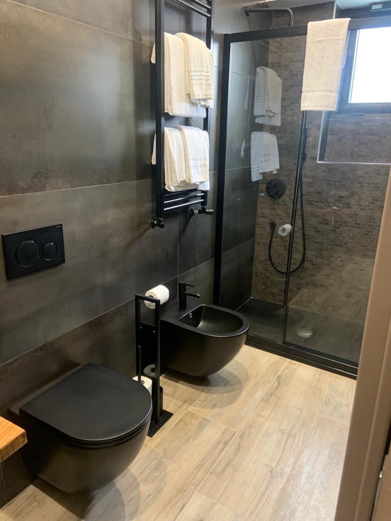 A modern bathroom with a black wall-mounted toilet, bidet, rainfall shower with glass enclosure, and heated towel rack with folded towels.