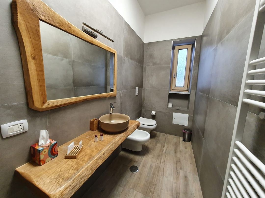 Modern bathroom featuring a wooden vanity with a gold vessel sink, large framed mirror, heated towel rack, and contemporary tile walls.
