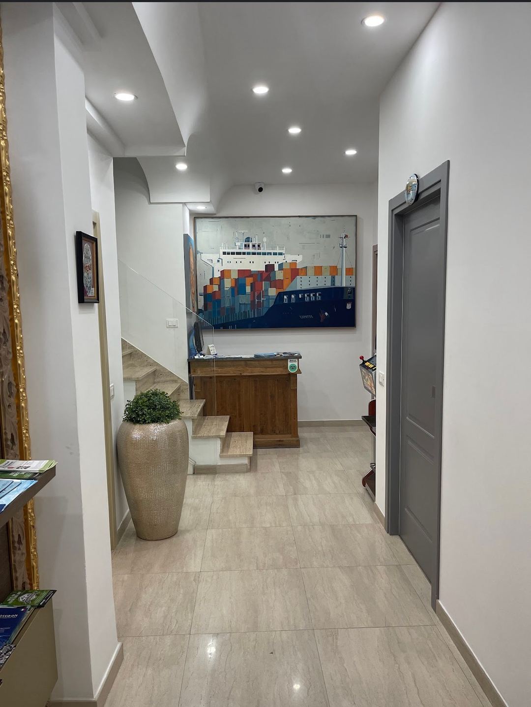 Modern hallway entrance of Villa Matilde B&B featuring white walls, recessed lighting, wooden furniture, and nautical-themed artwork of a cargo ship.